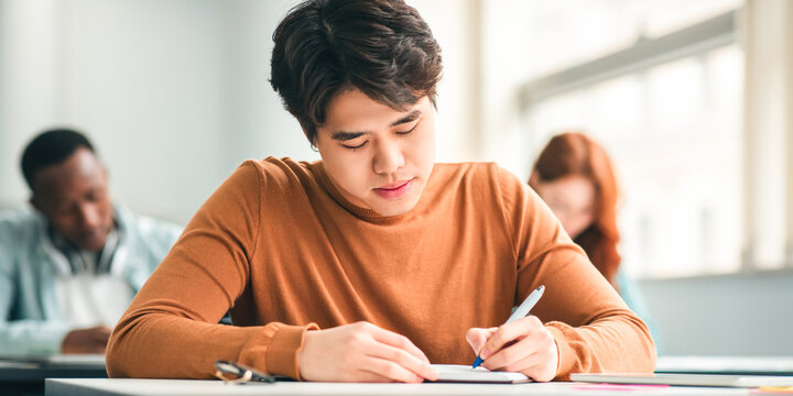 Education Concept. Portrait of serious asian male student sitting at desk in classroom at university, writing in notebook, taking notes, exam or test. Return to college and high school - Powered by Adobe