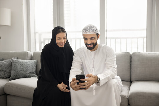 Connected in love. Happy smiling Arabic married couple dressed in religious Islamic tradition rest on comfy sofa at living room on weekend enjoy shopping online text messages watch video on smartphone - Powered by Adobe
