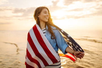 Happy woman on the beach with American flag on her shoulders. Beautiful woman enjoying sunset at sea, feeling freedom holding US flag. Independence and celebration concept.