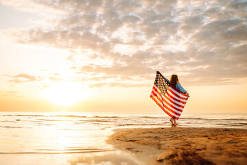 Happy woman on the beach with American flag on her shoulders. Beautiful woman enjoying sunset at sea, feeling freedom holding US flag. Independence and celebration concept.
