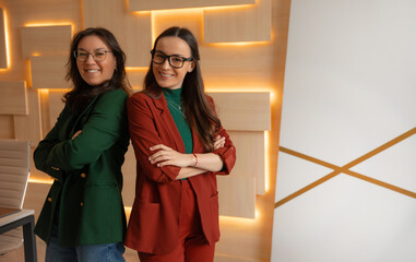 Two confident businesswomen stand back-to-back with arms crossed, smiling in a modern office....