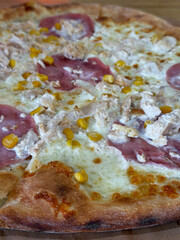 A close-up of a freshly baked pizza featuring a creamy white sauce, topped with chunks of chicken, slices of cured meat, and bright yellow corn kernels, highlighting its delicious textures.