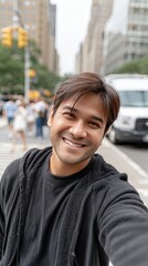 Fototapeta premium Asian man smiles at the camera while taking a selfie in City, capturing the lively atmosphere on a sunny day