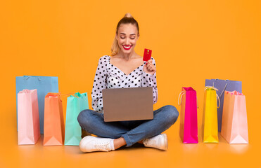 Online Payment Concept. Smiling young woman holding debit credit card and using laptop, ordering clothes and purchases via internet, sitting on the floor with colorful shopping bags at yellow studio