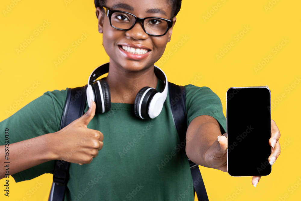 Poster Joyful Black Female Student Showing Smartphone With Empty Screen To Camera And Gesturing Thumbs Up Recommending Application For Cellphone Standing On Yellow Studio Background. I Like This App. Mockup