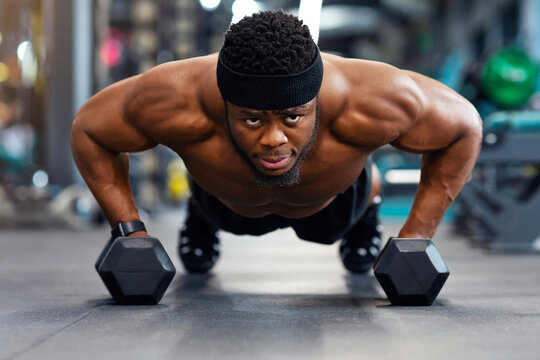 Confident black guy sportsman pushing up with dumbbells, looking at camera, panorama. Muscular african american shirtless man holding barbells and pushing from floor, work out at gym - Powered by Adobe