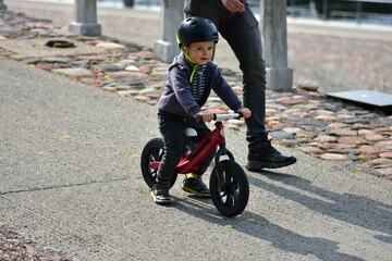 Young toddler boy riding a balance bike on a city footpath with his father. Child wearing helmet and sporty outfit enjoys outdoor fun and physical activity on a sunny day.