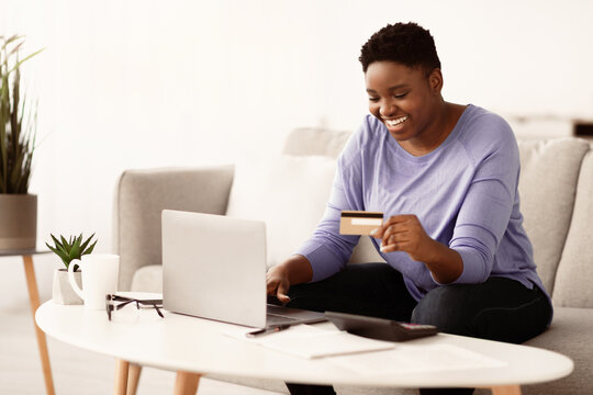 Easy Payment And Cashback Concept. Portrait of smiling african american young chubby woman holding credit card in hand and using laptop, sitting on couch at home, typing on keyboard, copy space