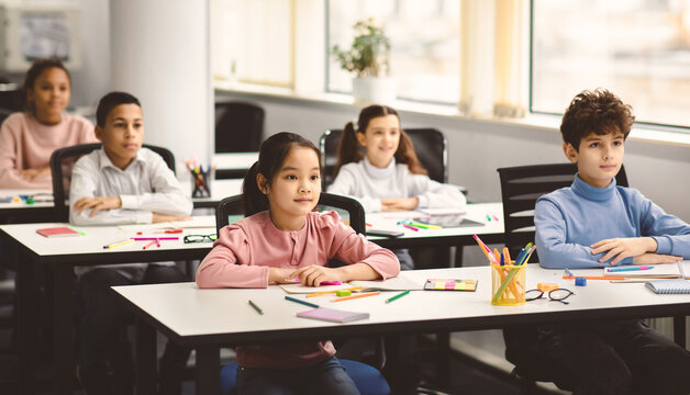 Back To School. Portrait of focused multicultiral pupils sitting with folded arms at desks with social distance, listening to teacher. Diverse junior students working diligently and learning new stuff - Powered by Adobe