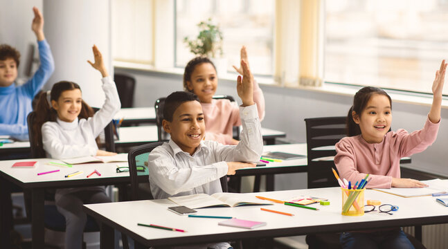 Ready For An Answer. Diverse group of multicultural cheerful primary schoolkids sitting at table in classroom and raising hands, studying and learning. Portrait of positive little elementary students - Powered by Adobe
