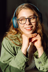Young woman in glasses listens to music on wireless headphones and enjoys the sun in a cozy...