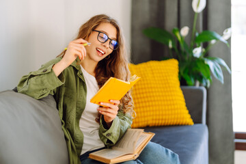 Portrait of smiling woman with yellow notebook in hands sitting on sofa in cozy room. Young female...