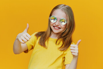 Cheerful girl with sunglasses giving thumbs up against a bright yellow background, expressing...