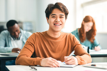 Modern Education Concept. Portrait of smiling asian male student sitting at desk in classroom at university, taking test or writing notes in his notebook, looking posing at camera, selective focus