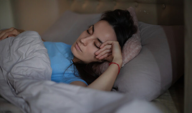 Young woman peacefully sleeping in bed, resting her head on a soft pink pillow with a gray blanket covering her. Cozy bedroom atmosphere, relaxation and nighttime rest concept.