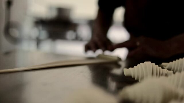 Close-up video of a baker in an apron preparing croissants from dough on a metal table in a professional kitchen. Hands carefully shape and roll the dough into croissants.