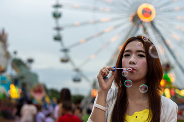 A young woman enjoying a day at the fair, blowing bubbles with a colorful ferris wheel in the background.