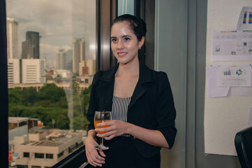 Professional woman holding a drink, dressed in a sleek outfit, with a city skyline backdrop, exuding confidence.