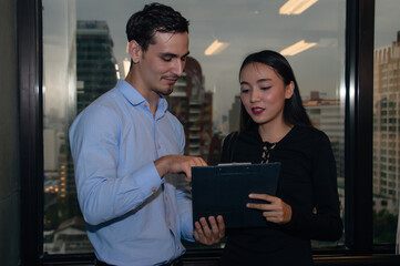Two professionals collaborating on a digital tablet, discussing ideas in a modern office setting with city skyline view.