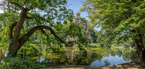 Paris, France - 06 14 2025: Buttes-Chaumont Park. Panoramic view of remarkable Sophora of Japan tree, belvedere island, Temple of the Sibyl, the lake and vegetation behind
