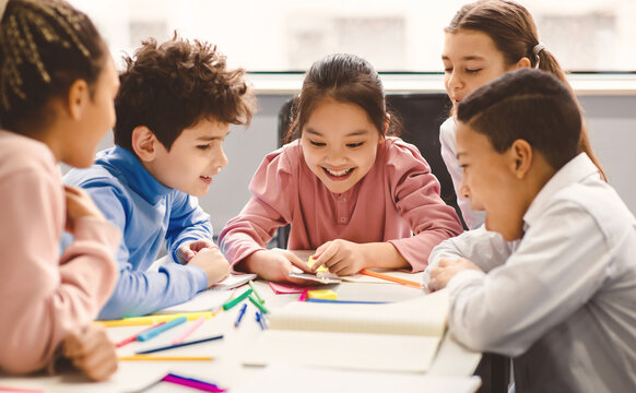 Technology And Pupils Concept. Group of excited multicultural happy junior children sitting at table and using smartphone, playing online mobile games. Modern Device, Gadget Addiction