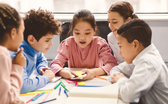 Diverse Group of Cute Small Children Using Smartphone, Sitting Together At Desk In Classroom Having Fun. Multicultural Kids Browsing Internet and Playing Online Video Games on Mobile Phone