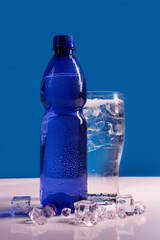 transparent glass of water with ice and blue bottle on light background