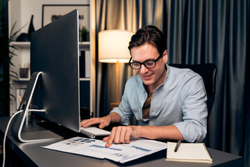 Smart businessman typing with keyboard on monitor with business paragraph dynamic data marketing analysis planning sheet aligning on report paperwork at modern home office at night time. Pecuniary.