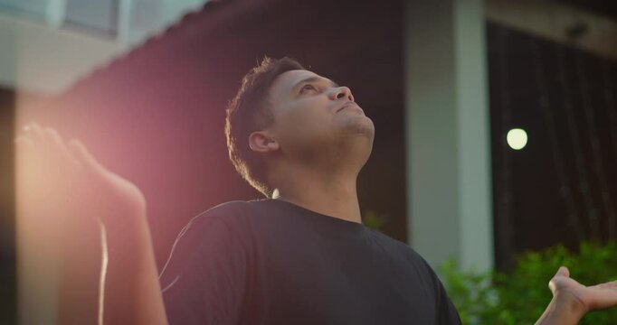 Latin American man praying with arms raised and head tilted upward, emotional gesture in outdoor evening light with greenery and building behind