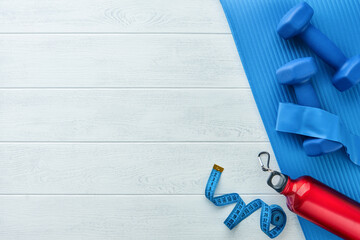 Fitness or healthy lifestyle concept. Blue dumbbells, red bottle, exercise mat and measuring tape on white wooden background with copy space. Minimal sports accessories idea. Top view with copy space