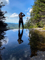 Woman and her reflection standing near small pond in forest