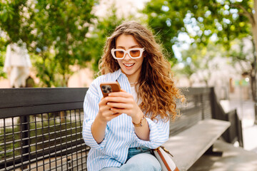Portrait of young woman with phone in hands enjoying walk in city. Cheerful woman sitting on bench...