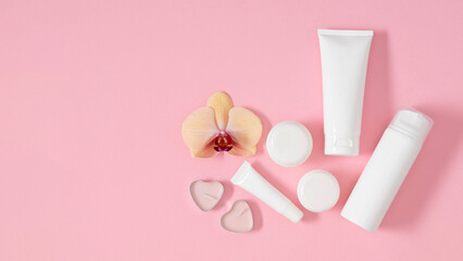 Cosmetic products in white plastic containers and bottles surrounded by candles and beautiful orchid on pink background on pink background. Fashionable composition of cosmetic products