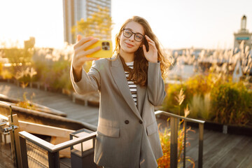 A beautiful woman enjoys the sunset and takes a selfie on her phone. A stylish business woman blogs...