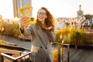 A beautiful woman enjoys the sunset and takes a selfie on her phone. A stylish business woman blogs...