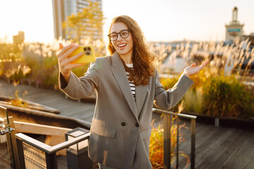 A beautiful woman enjoys the sunset and takes a selfie on her phone. A stylish business woman blogs...