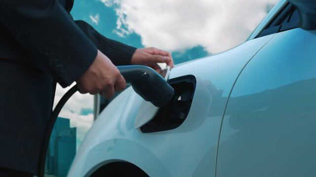 Electric vehicle, Businessman charges EV car battery energy at charging station on the freeway in urban area surrounded by tall skyscrapers. Eco friendly progressive clean energy concept.