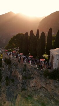 Aerial view of luxury wedding at Villa Cimbrone, Ravello. Guests dance on a cliff-edge terrace above a sheer stone wall, surrounded by red umbrellas, sea haze, and golden Amalfi Coast sunset
