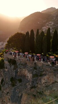 Aerial view of luxury wedding at Villa Cimbrone, Ravello. Guests dance on a cliff-edge terrace above a sheer stone wall, surrounded by red umbrellas, sea haze, and golden Amalfi Coast sunset