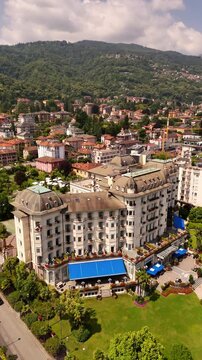 Drone video of Stresa town on Lake Maggiore, Italy, featuring historic grand hotels, bright midday sun, and cars driving along roads near the lake. Vibrant lakeside scene
