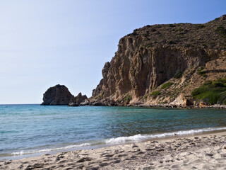 Sunlit beach with gentle waves, warm sand, and stone cliffs — a typical Greek summer landscape.