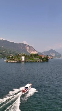 Drone view of a private wooden luxury yacht sailing on Lake Maggiore toward Isola Bella, featuring lush gardens and a majestic baroque palace. Iconic destination for exclusive travel in Northern Italy
