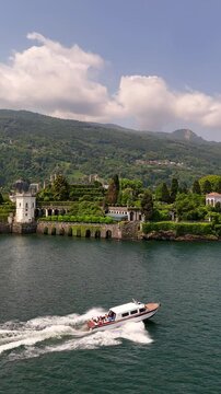 Drone view of a private wooden luxury yacht sailing on Lake Maggiore toward Isola Bella, featuring lush gardens and a majestic baroque palace. Iconic destination for exclusive travel in Northern Italy