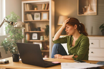 A woman appears overwhelmed while seated at her home office desk, glancing down at her laptop in...