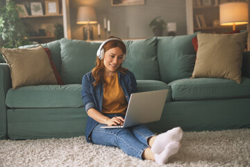 A young woman, wearing headphones, is seated on the floor with her laptop, focused on her work in a...