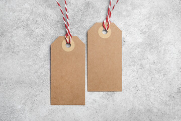 Two vertical blank kraft paper tags on red and white rope on a textured light gray concrete...