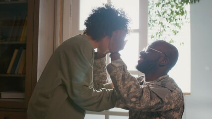 Medium closeup of cheerful black male soldier in khaki military uniform arriving home from overseas tour of duty, lovingly hugging young son, patting him on head and admiring