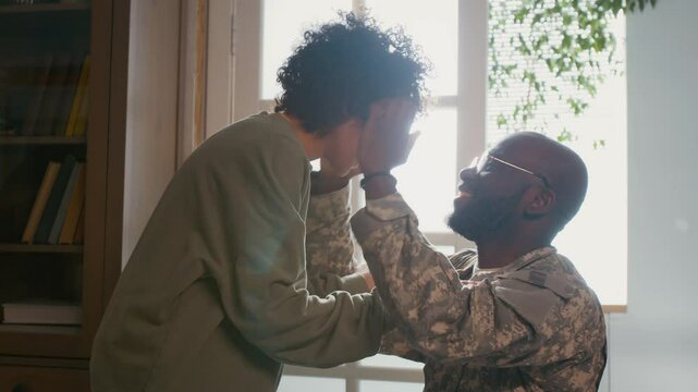 Medium closeup of cheerful black male soldier in khaki military uniform arriving home from overseas tour of duty, lovingly hugging young son, patting him on head and admiring