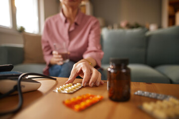 A senior woman is selecting medication from a blister pack on a table, with a glass of water nearby, while sitting comfortably on a couch in her living room.