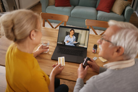 An elderly couple sits at their table, holding medications while talking to a doctor on a laptop during a telemedicine appointment at their home.
